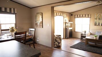 A spacious living area with a dining table, chairs, a sofa, and a coffee table. There is a large window providing natural light, and a door leading to another room.