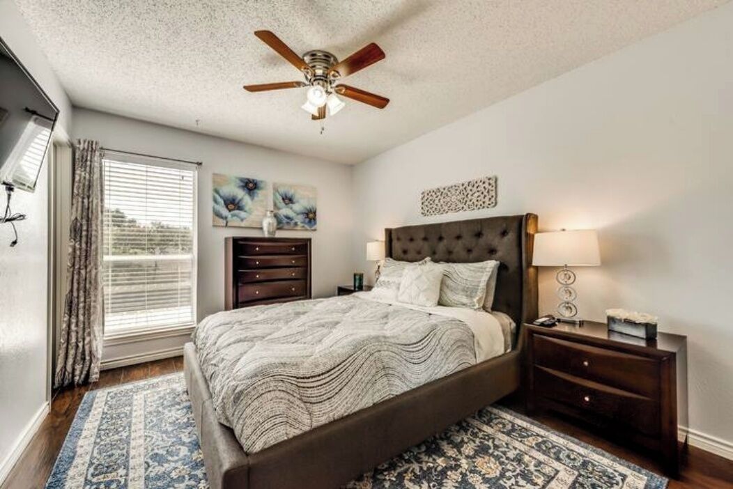 A bedroom with a bed, a ceiling fan, a nightstand, a dresser, and a window with curtains.
