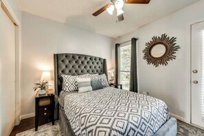 Une chambre à coucher avec une tête de lit capitonnée, une couvre-lit à motifs, une table de chevet avec une lampe et un miroir décoratif accroché au mur.