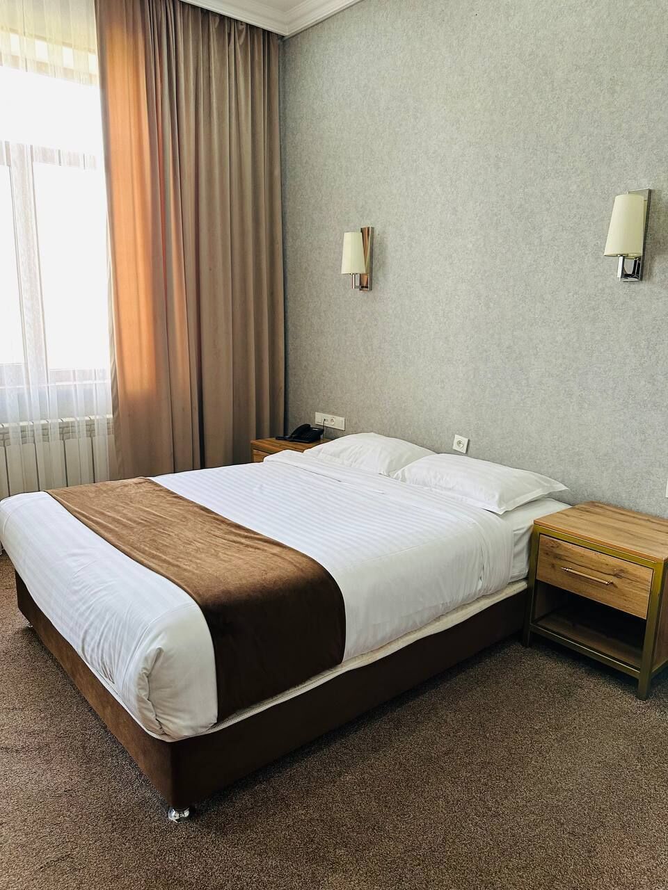 A hotel room with a large bed, a nightstand, a lamp, and a view of the outdoors.