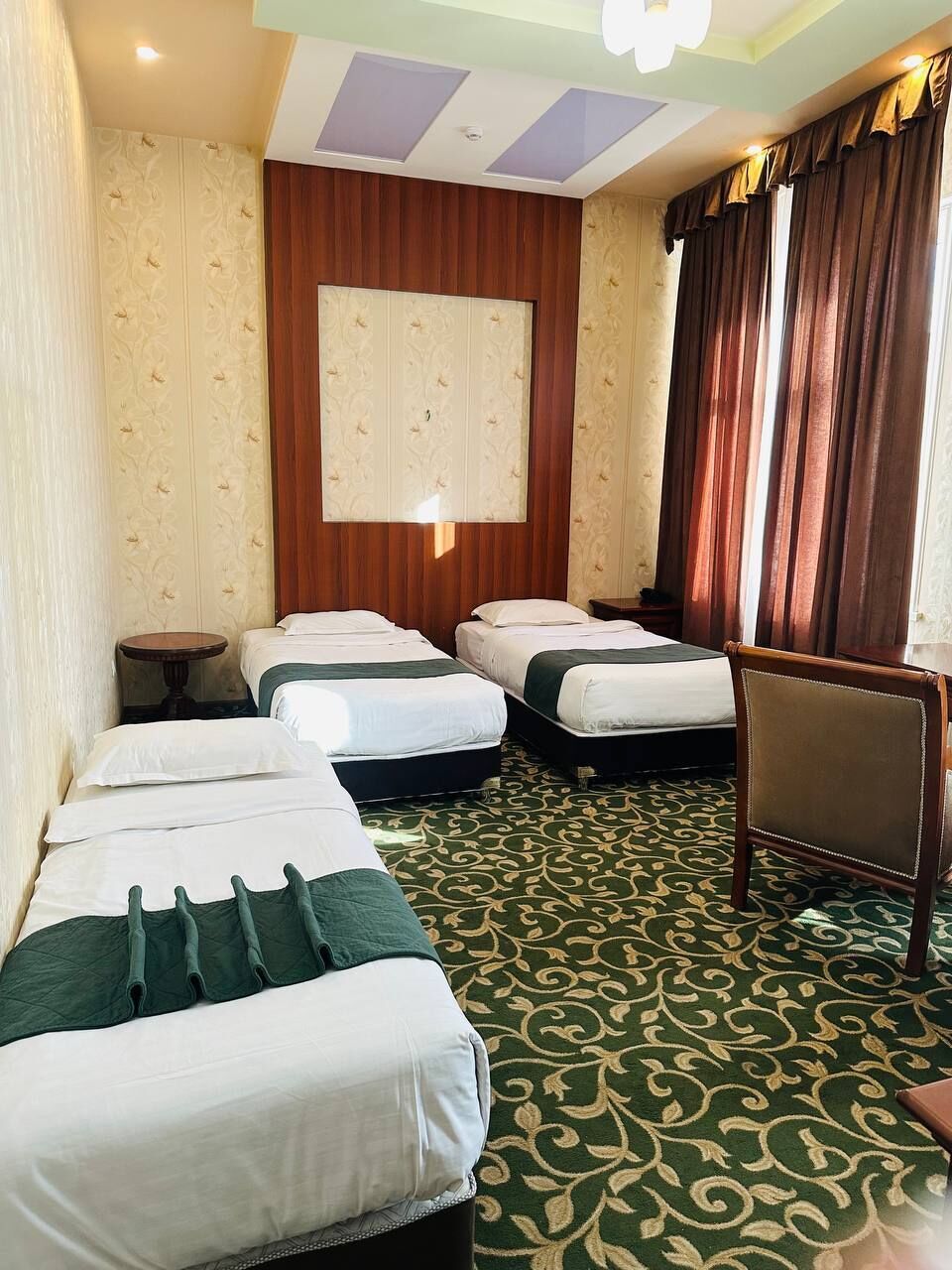 Une chambre d’hôtel avec trois lits, une table ronde, une chaise, une grande fenêtre avec des rideaux et un tapis à motifs.