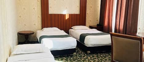 Une chambre d’hôtel avec trois lits, une table ronde, une chaise, une grande fenêtre avec des rideaux et un tapis à motifs.