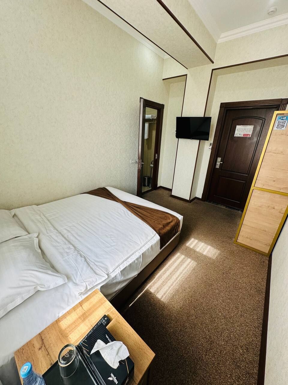 A hotel room with a single bed, a wooden nightstand, a television, and a door with a sign.