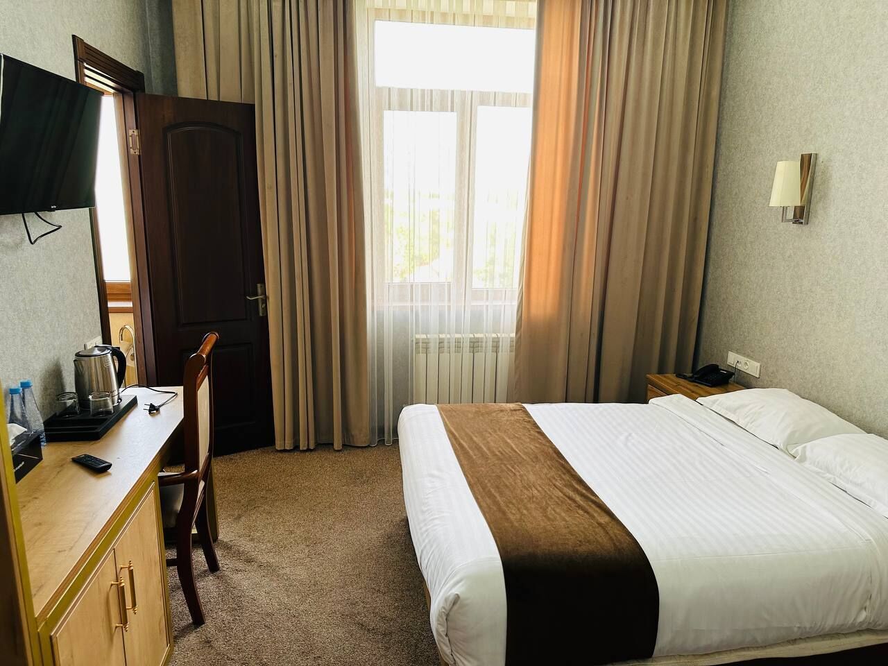 A hotel room with a bed, a desk with a telephone, a chair, a television, and a window with curtains.