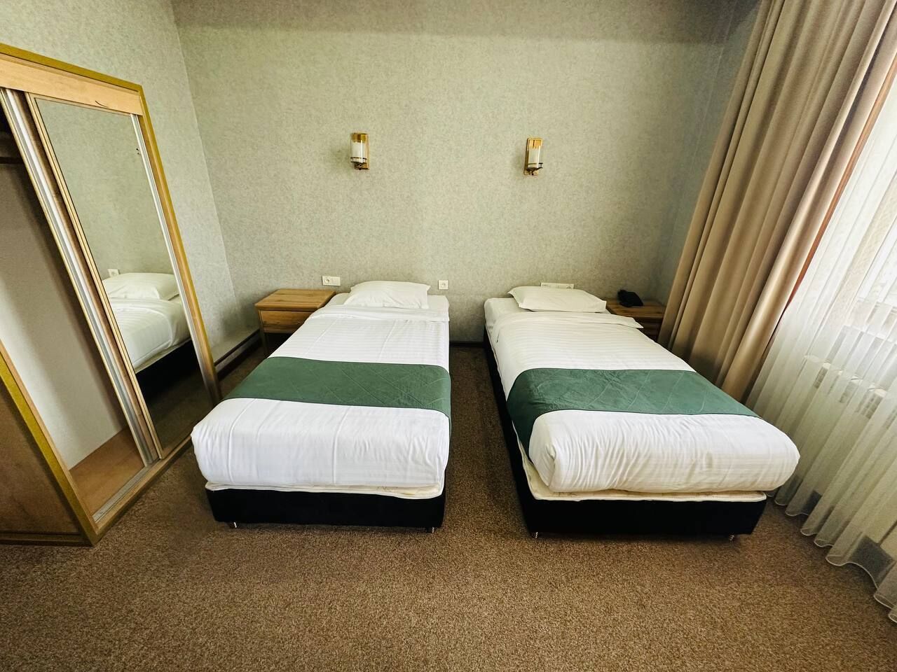 A hotel room with two single beds, a large mirror, bedside tables, and a window with curtains.