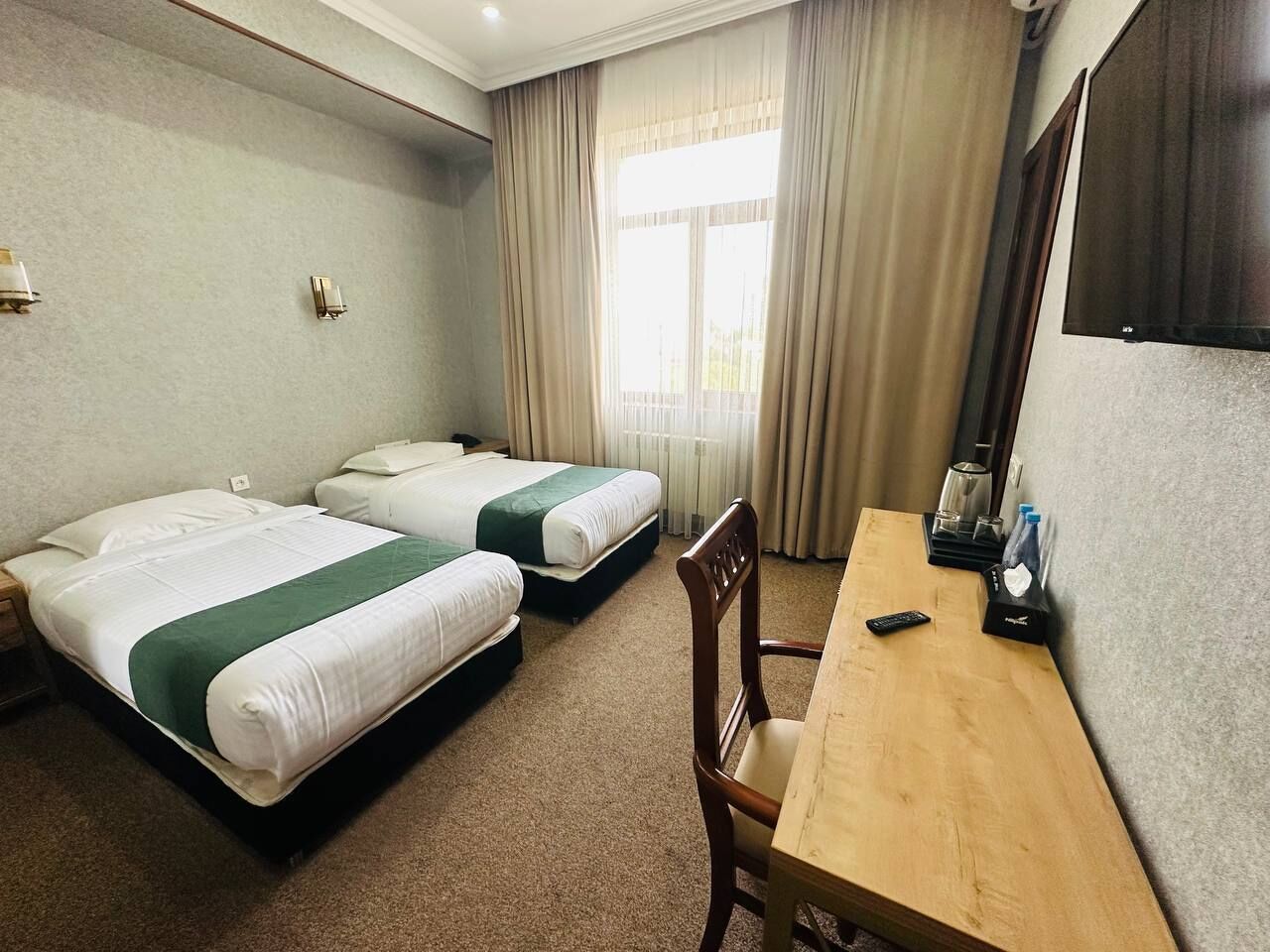A hotel room with two beds, a wooden desk, a chair, a TV, and a window with curtains.