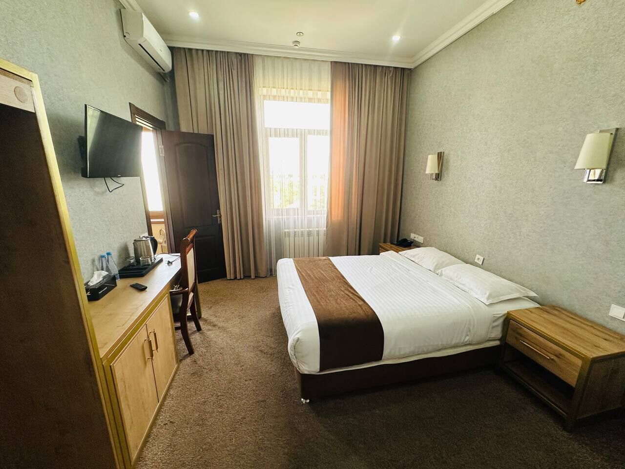 A hotel room with a bed, bedside table, desk with a chair, a television, and a window with curtains.