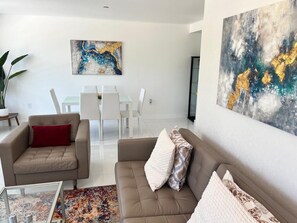 A modern living room with a beige sofa, a red pillow, and a patterned rug. There is a dining area with white chairs and a table in the background.