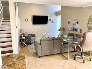 Un salon moderne avec un canapé gris, une table basse en verre, un téléviseur fixé au mur et un escalier menant à un autre niveau.