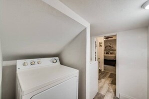 A laundry room with a front-loading washer and dryer, a wooden herringbone floor, and a doorway leading to a bedroom with a bed and a nightstand.