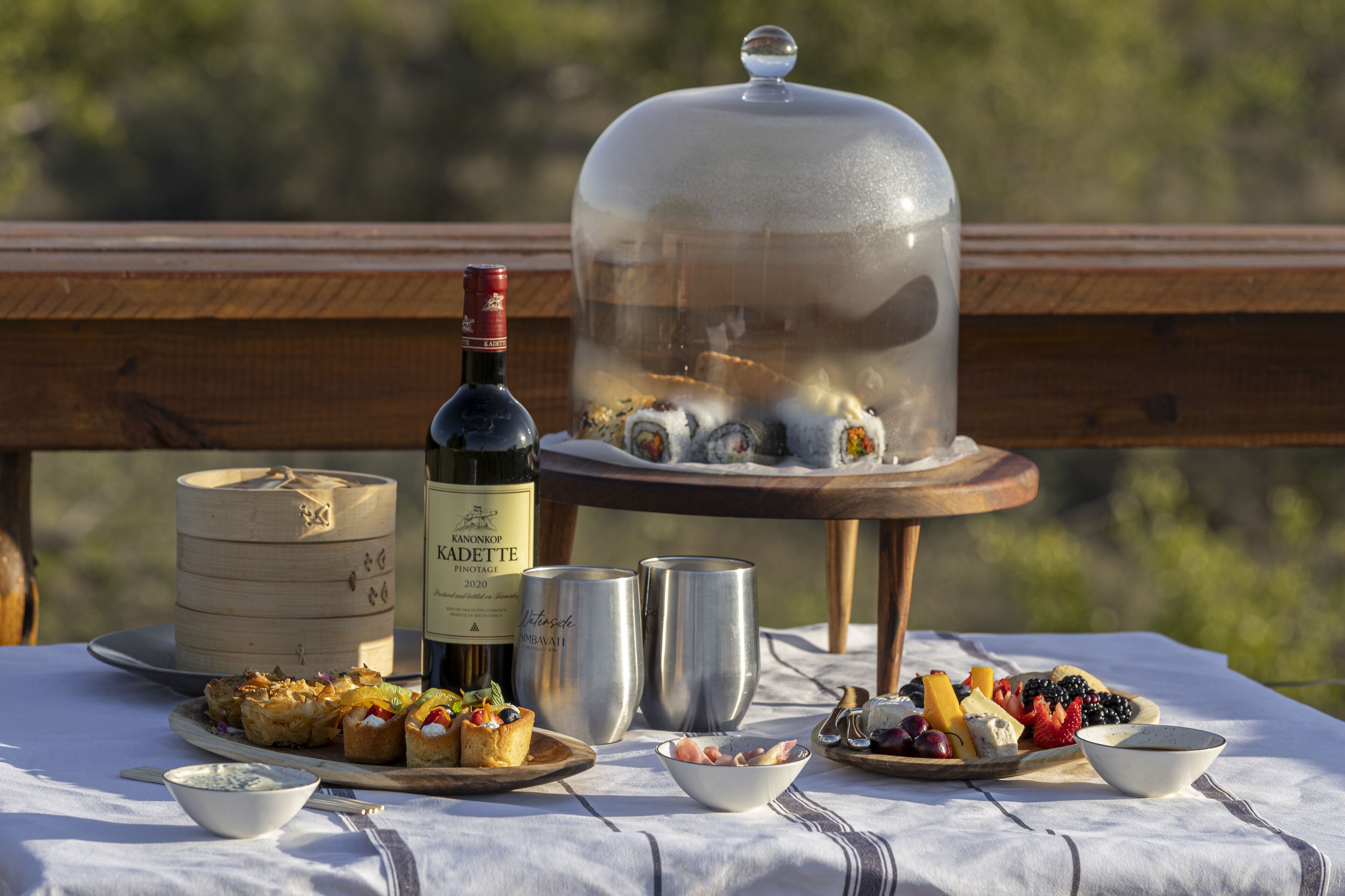Una mesa dispuesta con comida y bebidas, incluyendo una botella de vino, un recipiente de madera y una cúpula de vidrio que cubre sushi.