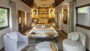 Un dormitorio amplio con una cama grande, dos sillones, un otoman redondo y un tocador con espejo e iluminación.