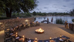 Un espacio al aire libre con mesas y sillas dispuestas alrededor de una chimenea central, con vista a un cuerpo de agua.