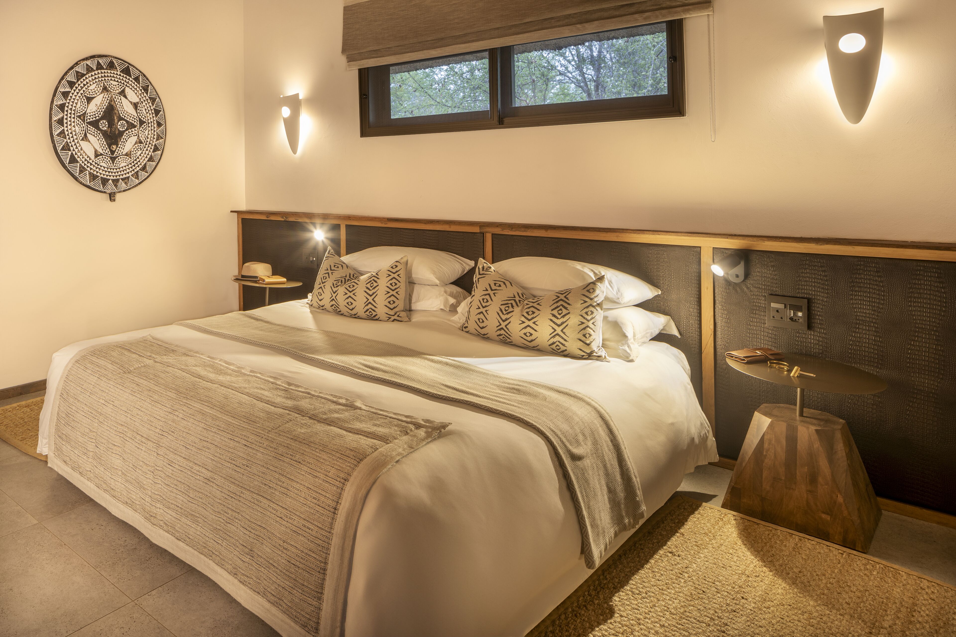 Een netjes ingerichte slaapkamer met een groot bed, bedkussens met een patroon, een nachtkastje en een wandlamp.