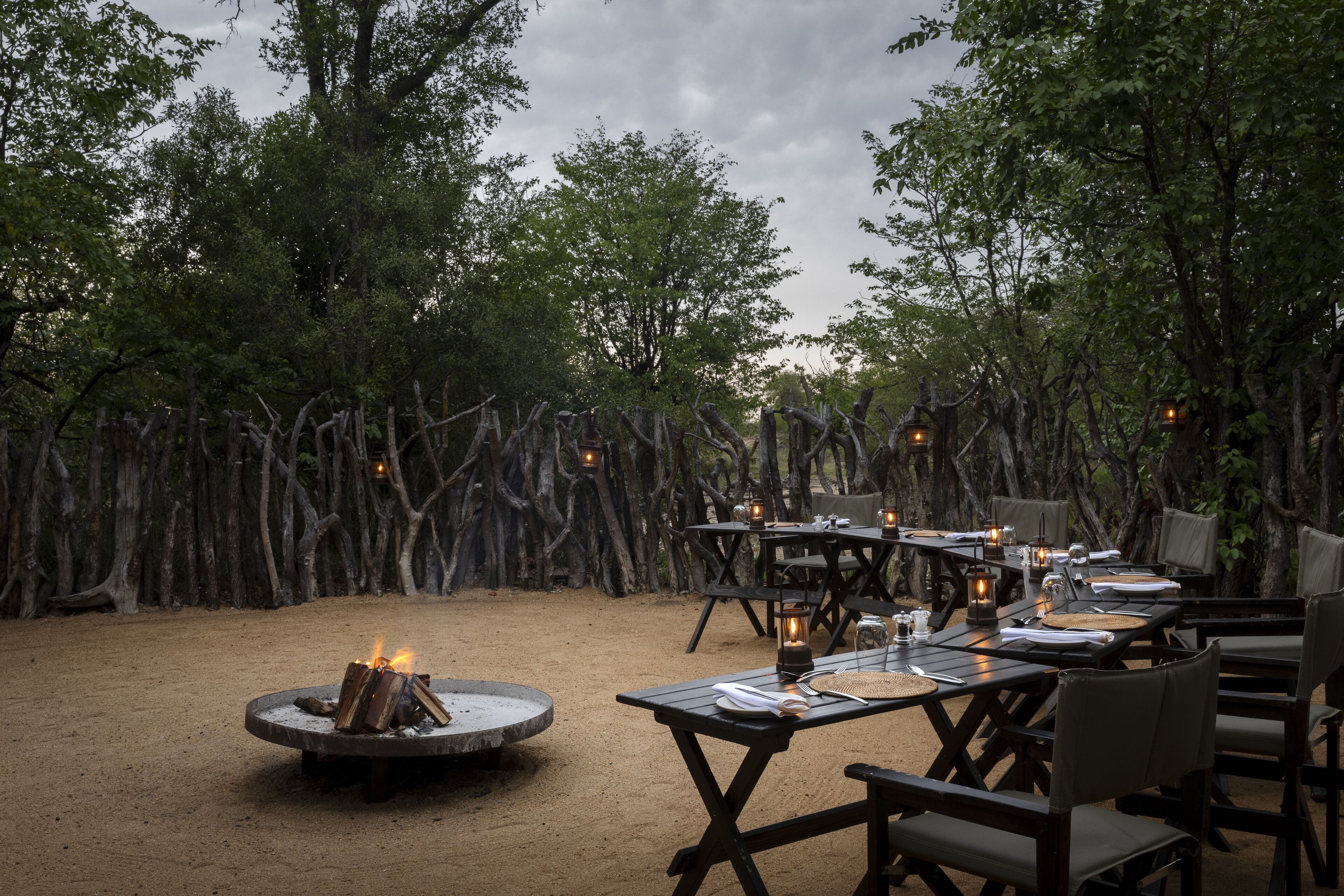 Un espace extérieur aménagé pour les repas, avec des tables et des chaises, entouré d’arbres et doté d’un foyer.
