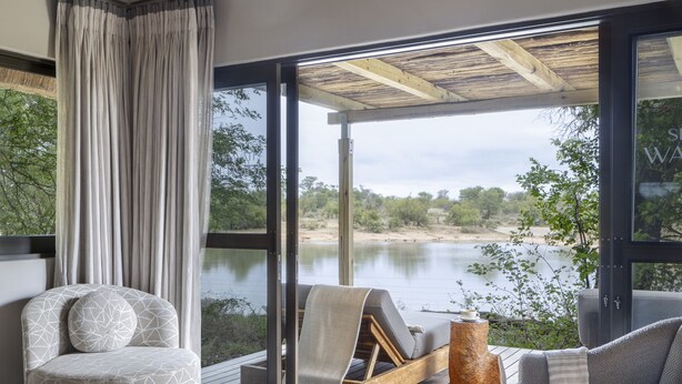 Un dormitorio moderno con una cama grande, dos sillones grises, un otoman redondo y vistas a un lago a través de amplios ventanales.