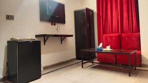 A room with a flat-screen TV mounted on the wall, a small table with a chair, a red sofa, and a black refrigerator. - Hotel Royal Grand (Mekhtar)