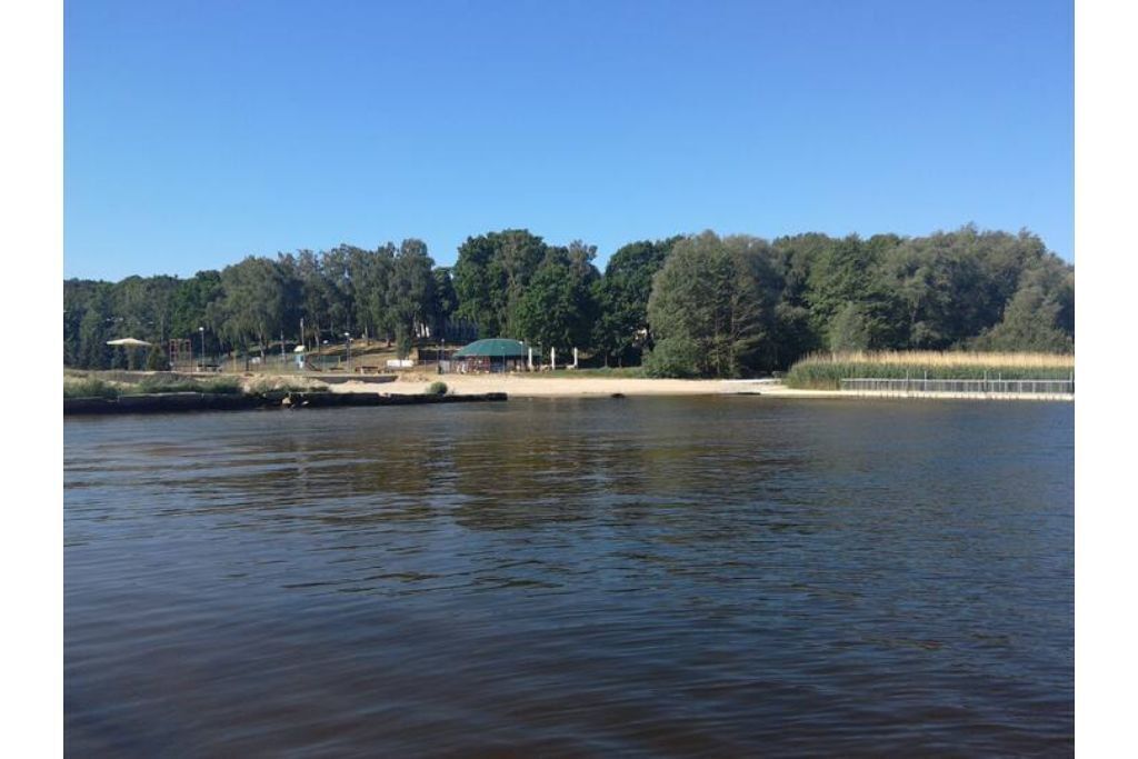 A calm lake with a sandy shore, surrounded by dense trees and a small pavilion.