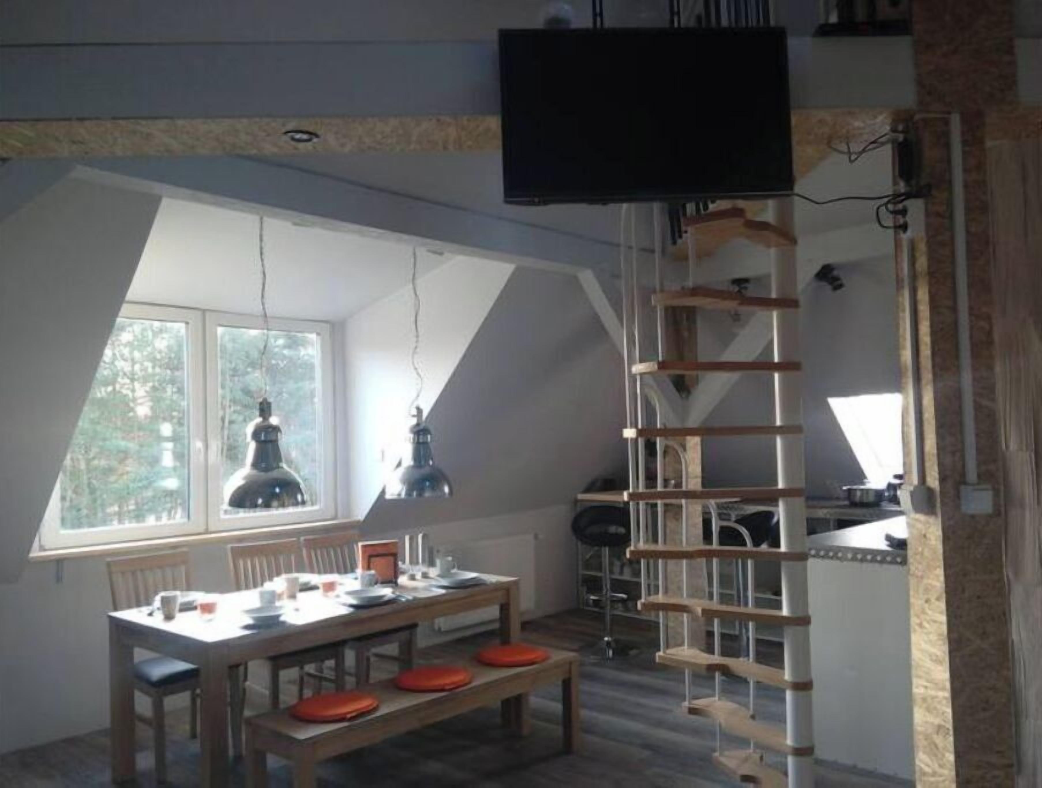Een eetruimte met een hoge plafond, een houten tafel en banken. Er hangt een televisie aan de muur en er staat een ladder in de hoek.
