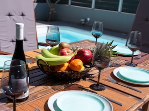A dining table with a fruit basket, wine glasses, and plates by a poolside.
