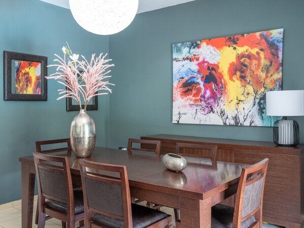 A dining area with a wooden table, six chairs, a vase with pink flowers, a large abstract painting, and a lamp.