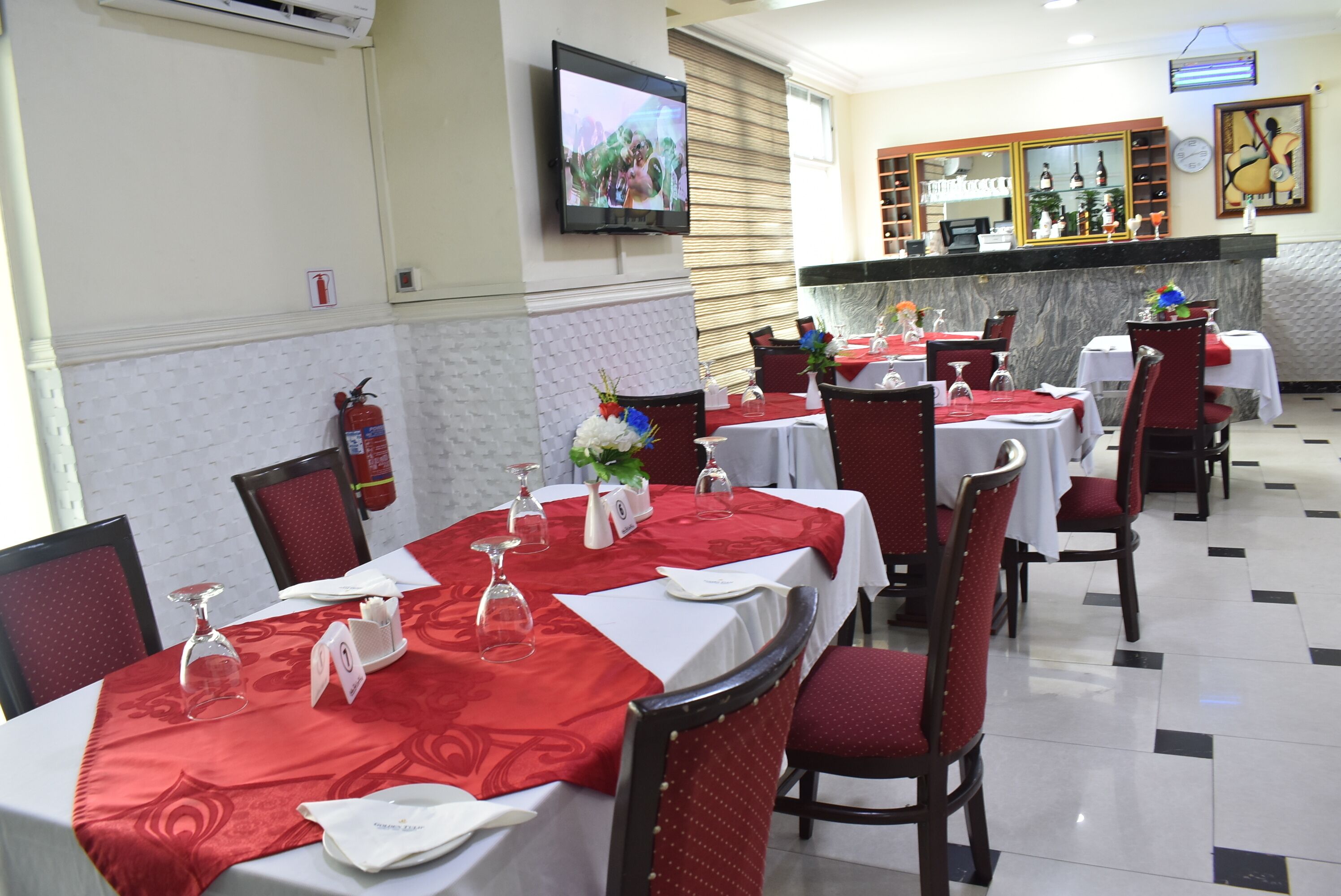Un restaurante con mesas dispuestas para comer, un extintor, un televisor montado en la pared y una barra con un espejo y elementos decorativos.