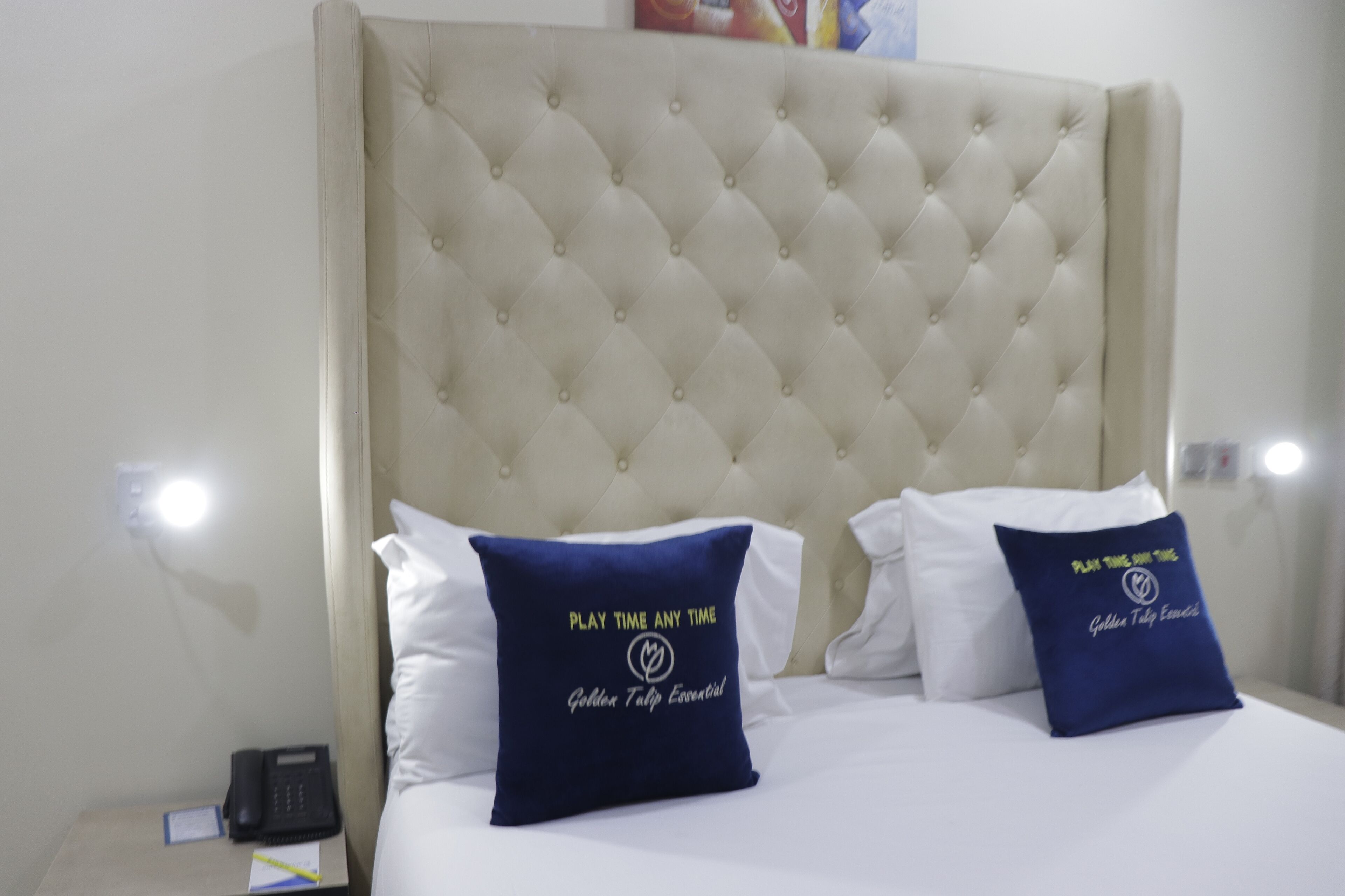 Una cama con cabecera capitonada, dos almohadas y una mesita de noche con un teléfono y un bloc de notas.