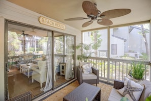 Une terrasse couverte, meublée de meubles en osier, avec un ventilateur de plafond et une enseigne indiquant « Maison de plage ».