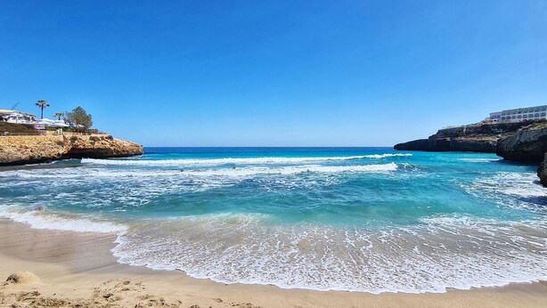 A beach with clear blue waters, rocky cliffs, and a building with multiple balconies overlooking the sea.