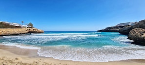 Una spiaggia con acque cristalline, scogliere rocciose e un edificio con diversi balconi affacciati sul mare.
