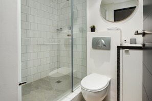 A modern bathroom with a glass-enclosed shower, a white toilet, a round mirror, and a small potted plant.