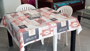 Un coin repas avec une table recouverte d’une nappe à motifs, deux chaises blanches, un canapé marron et un réfrigérateur en arrière-plan.