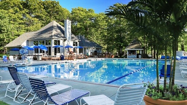 A swimming pool with lounge chairs and umbrellas, surrounded by trees and a building with a sloped roof. - Cozy Ski Condo with Pond View (Hidden Valley)