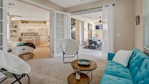 A spacious living area with a blue sofa, a white armchair, and a round coffee table. There is a kitchen in the background with white cabinets and a dining area visible through an open door. - Heron's Glen Beautiful Beachy Home in Gated Golf Community Home (North Fort Myers)