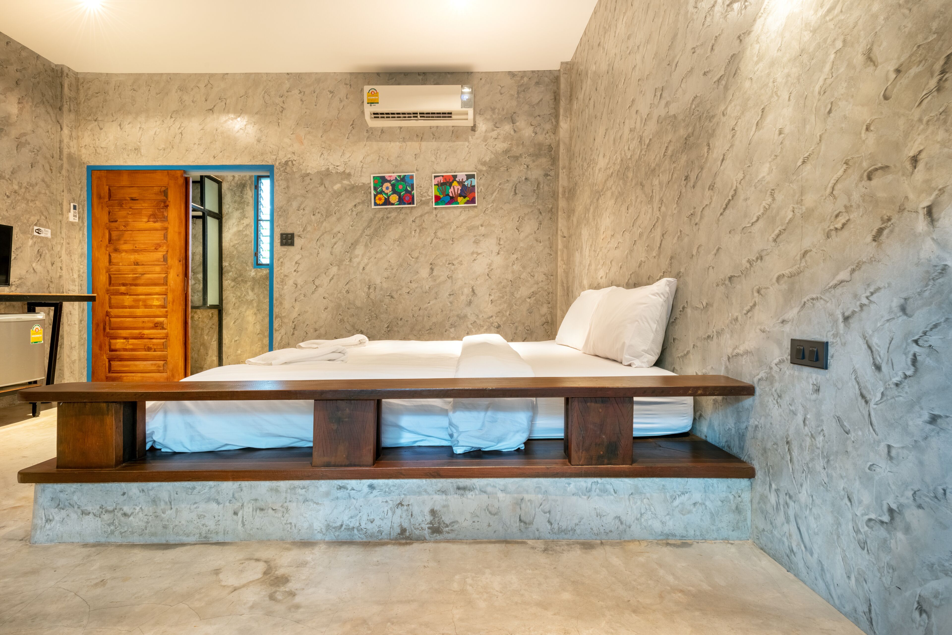A bedroom with a bed, a wooden bench, a door, and a wall-mounted air conditioner.
