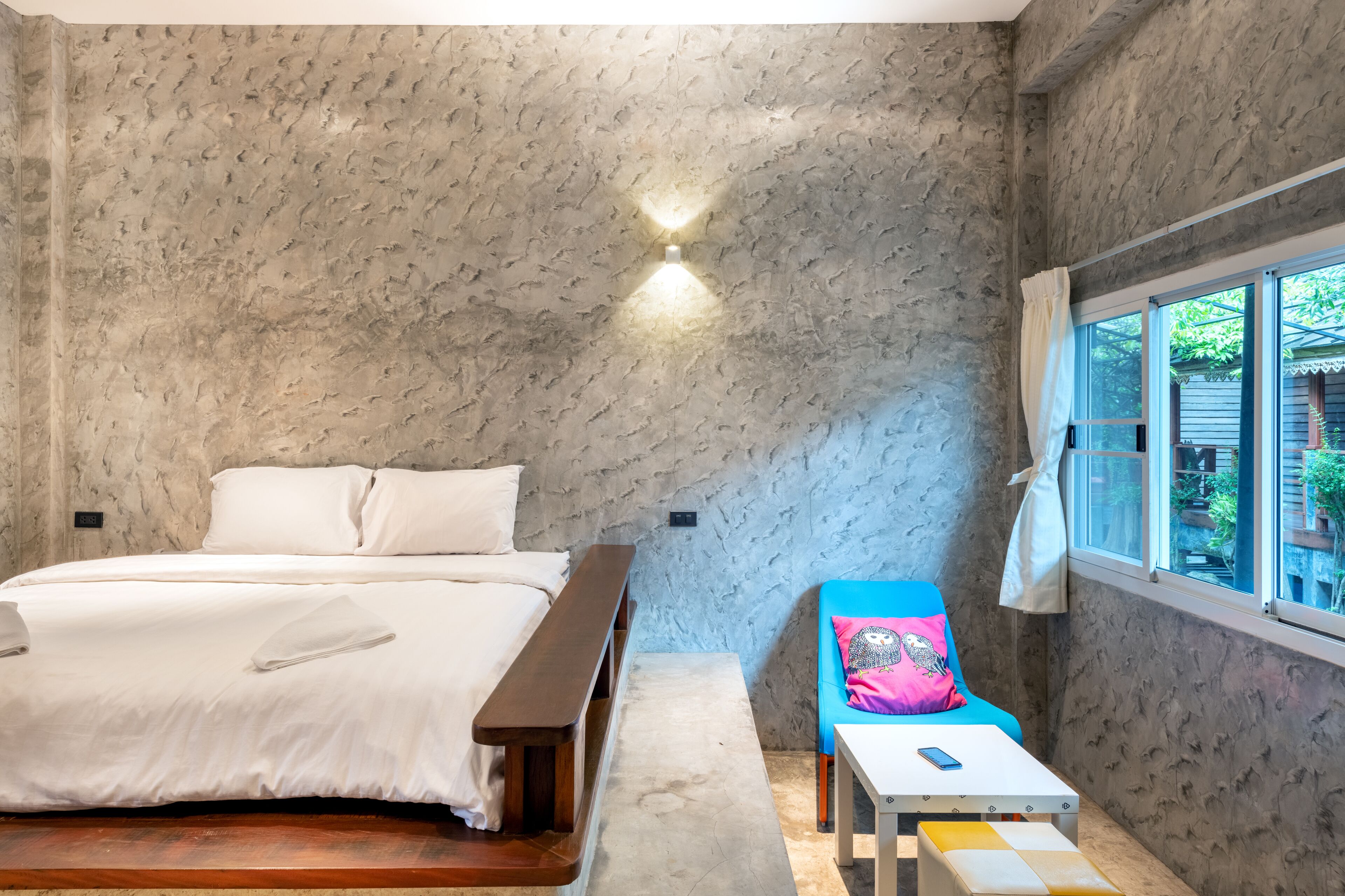 Un dormitorio con cama, un banco, una mesita y ventana con vistas a la vegetación.