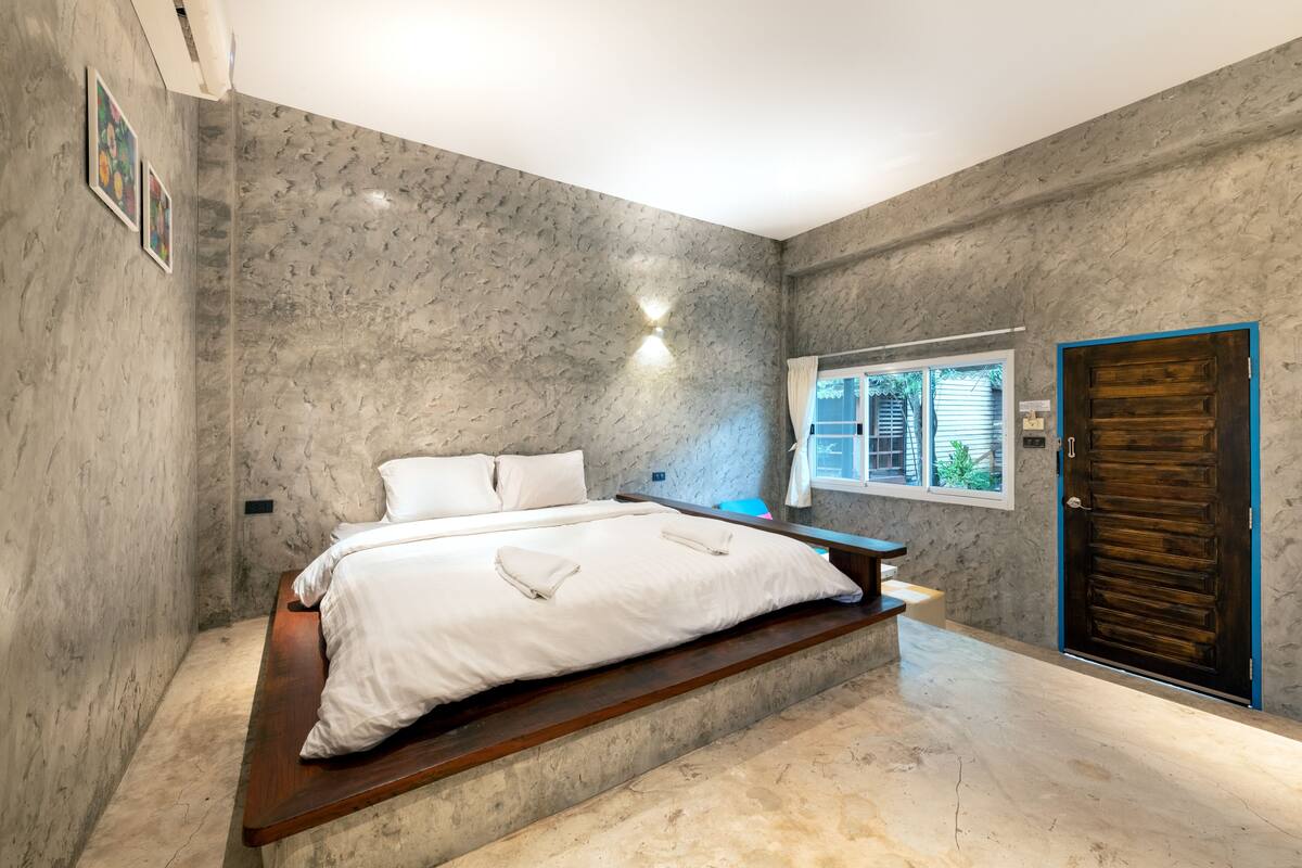 A bedroom with a bed on a wooden platform, a window with curtains, a door, and a textured wall.
