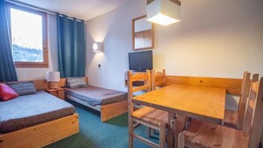 Una habitación pequeña y acogedora con dos camas de madera, una mesa de madera con sillas, un televisor y una ventana con cortinas.