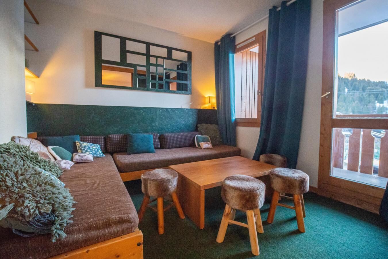 A cozy room with a sofa, a wooden table, and stools. There is a window with curtains and a view of the outside.