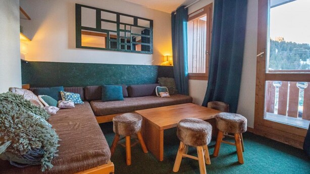 Una habitación acogedora con un sofá, una mesa de madera y taburetes. Se ve una ventana con cortinas y vistas al exterior.