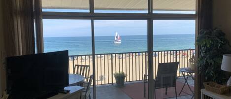 A room with a large sliding glass door leading to a balcony with a view of the beach and ocean.