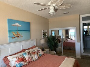 A bedroom with a bed, a ceiling fan, a painting of beach chairs, a mirror, and a door leading to another room. - Large Oceanfront condo overlooking the boardwalk.<br>Regency Unit 15 (Ocean City)