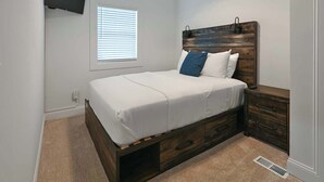 A bedroom with a bed, a nightstand, a wooden headboard, a TV mounted on the wall, and a window with blinds.