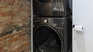 A compact, black wall-mounted washer and dryer unit in a bathroom with a brick wall and a white toilet.