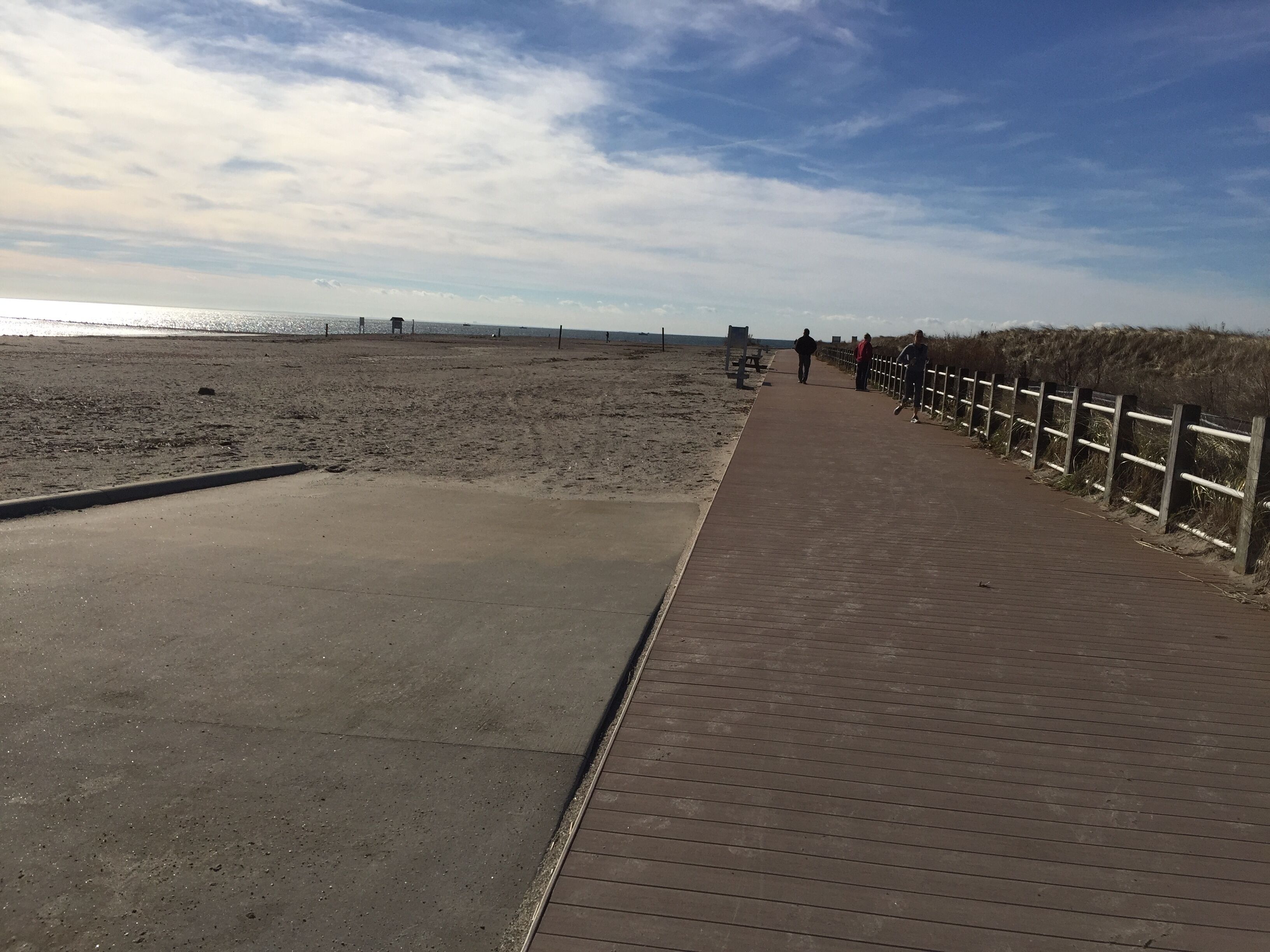 Une promenade en bois mène à une plage de sable, avec une clôture d’un côté et un chemin en béton de l’autre.