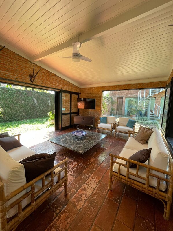 A spacious living area with a brick wall, a ceiling fan, a flat-screen TV, and a view of the garden through large glass doors. - House located 30 meters from Pernambuco Beach (Guarujá)