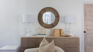 Una habitación con un aparador de madera, dos lámparas blancas, un espejo redondo y una pequeña mesa con adornos.