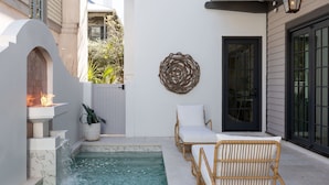 A small outdoor pool area with a fire feature, wicker chairs, and a decorative wall piece.