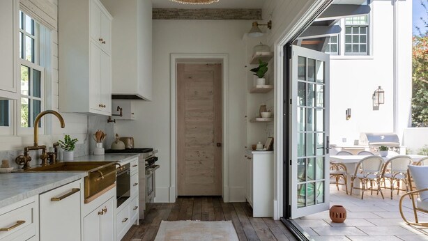 A modern kitchen with white cabinetry, a wooden door, and a view of an outdoor patio with wicker furniture.