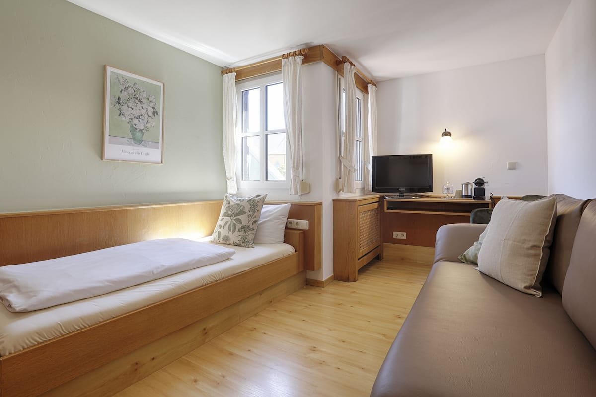 Um quarto aconchegante com uma cama, um sofá, uma escrivaninha com TV e uma janela com cortinas.